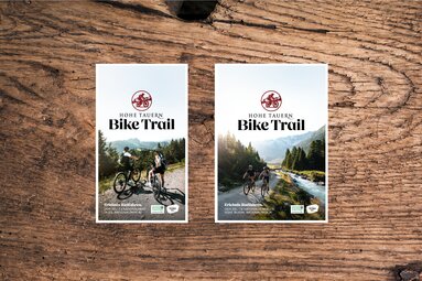 Zwei Hohe Tauern Bike Trail Titelbilder liegen nebeneinander auf Holz. | © Ferienregion Nationalpark Hohe Tauern