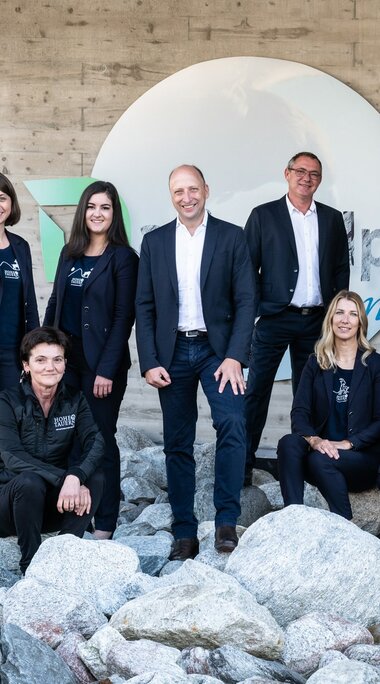 Team in Businesskleidung posiert auf Steinen vor modernem Gebäude mit rundem Logo. | © Foto Christine