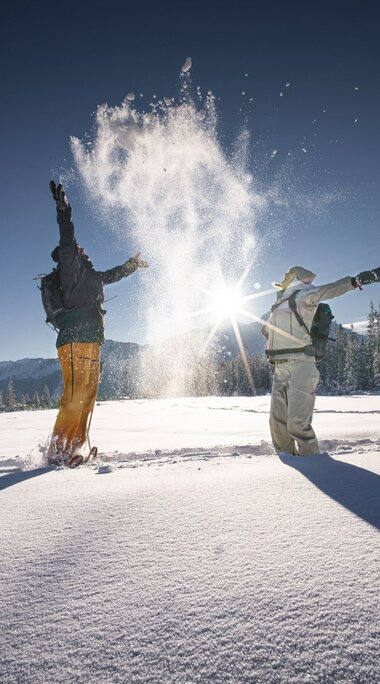 Zwei Menschen werfen Schnee in die Luft im Winterwald bei Sonne. | © Ferienregion Nationalpark Hohe Tauern - Daniel Breuer